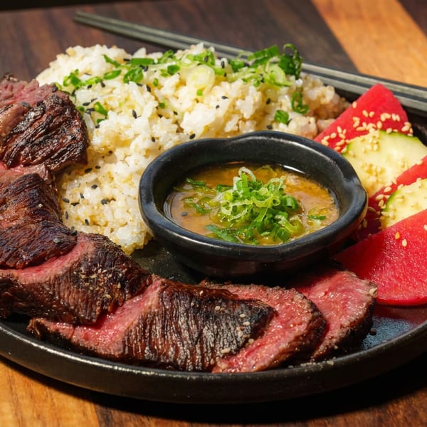 Sliced medium-rare steak, sesame rice, dipping sauce, watermelon, and cucumber on a black plate with chopsticks.