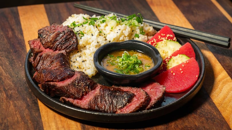 Sliced medium-rare steak, sesame rice, dipping sauce, watermelon, and cucumber on a black plate with chopsticks.