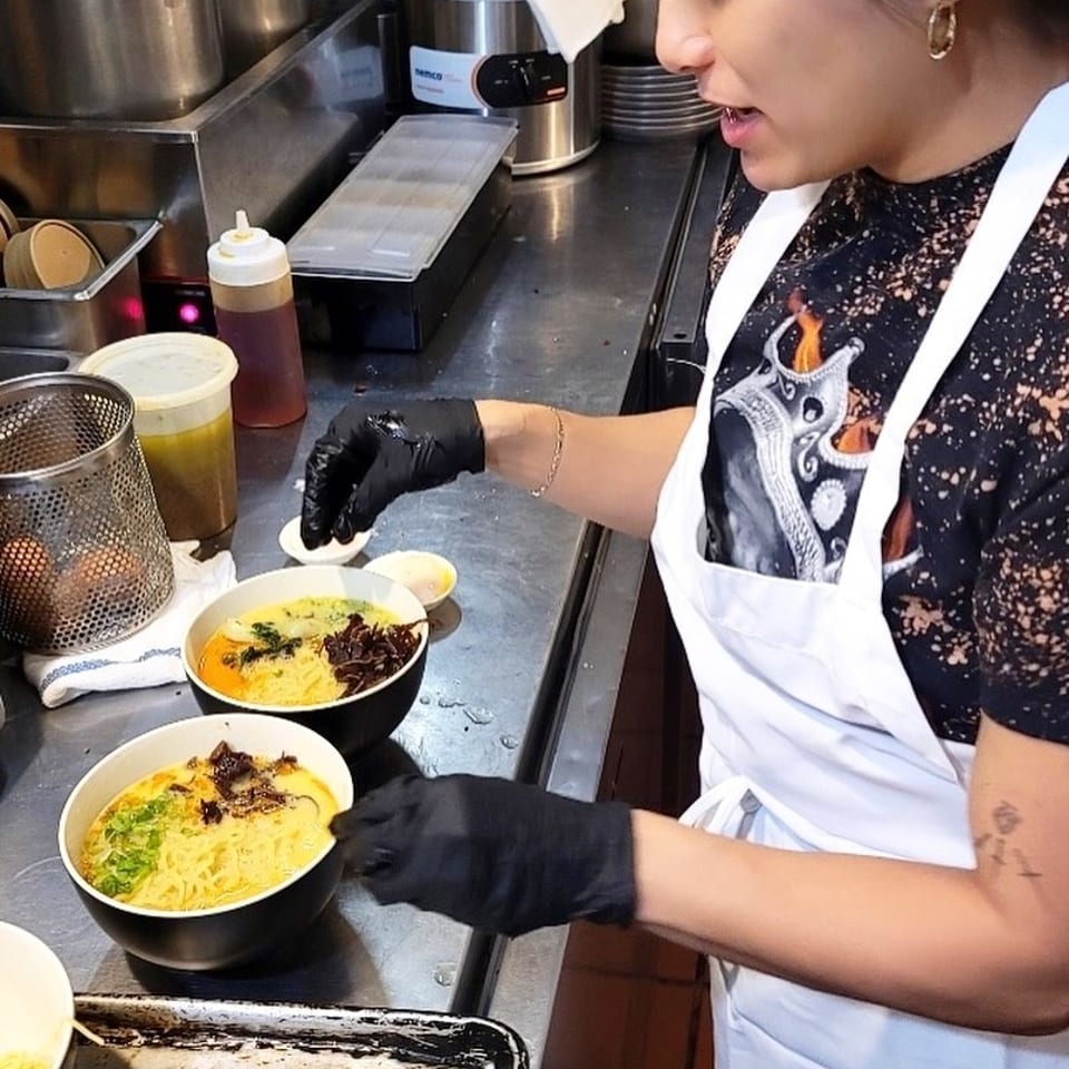 Chef wearing an apron and black gloves garnishes two bowls of ramen in a kitchen.