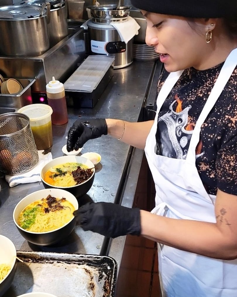 Chef wearing an apron and black gloves garnishes two bowls of ramen in a kitchen.