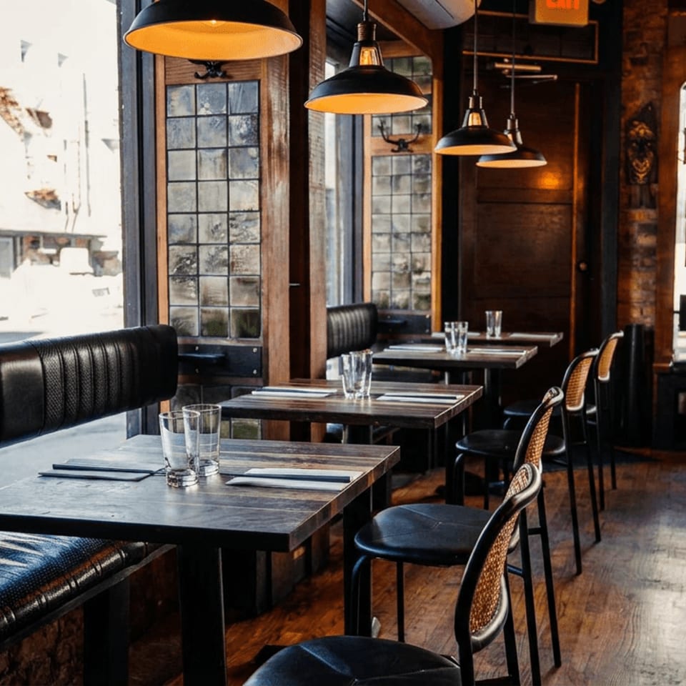 Cozy restaurant interior with wooden tables, black booths, cane chairs, and pendant lights by large windows.