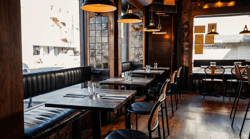 Cozy restaurant interior with wooden tables, black booths, cane chairs, and pendant lights by large windows.