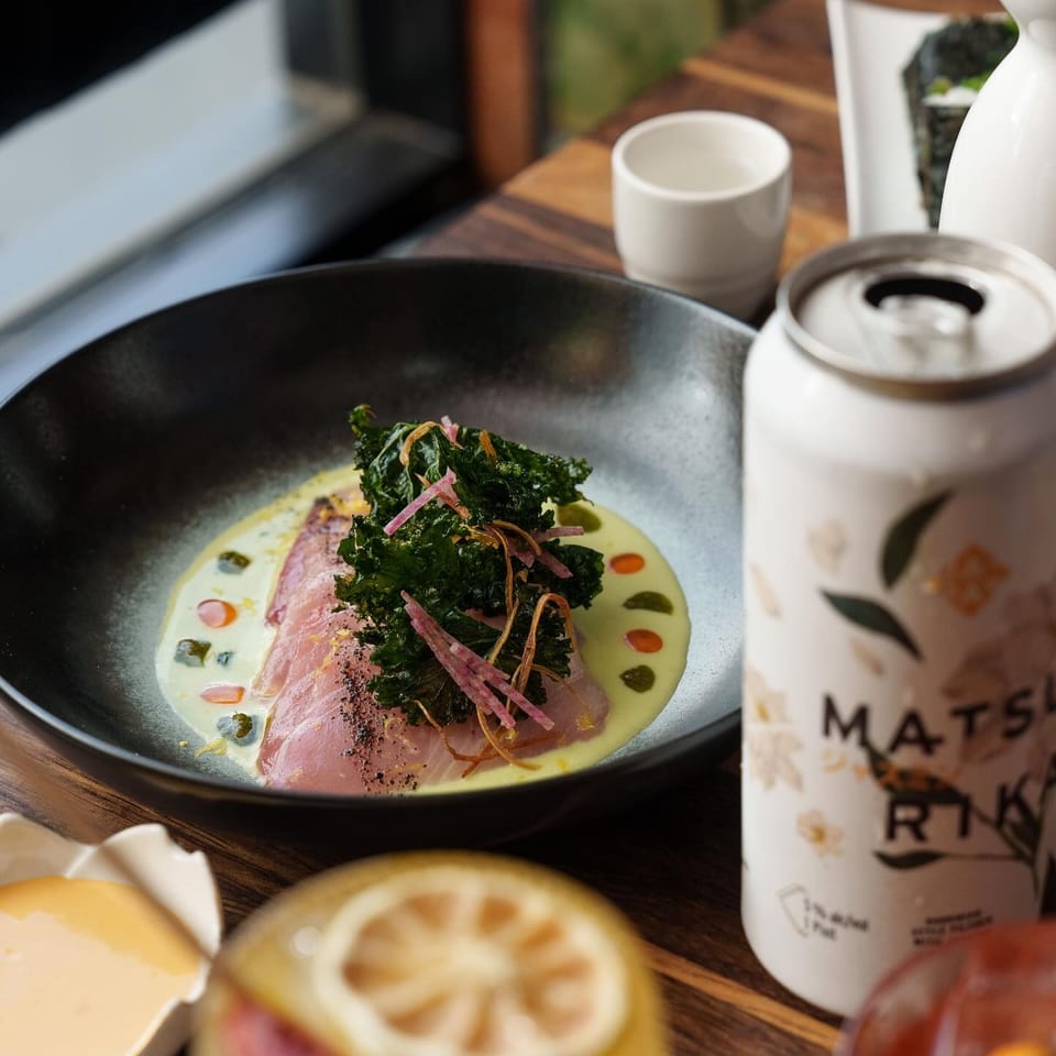 Sliced fish with kale and sauce in a black bowl, a Matsu Rika can, and cocktails on a wooden table.