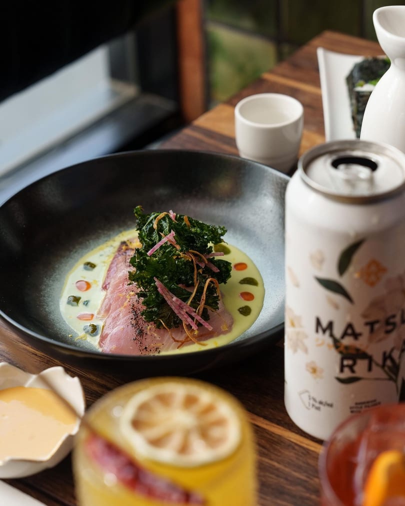 Sliced fish with kale and sauce in a black bowl, a Matsu Rika can, and cocktails on a wooden table.