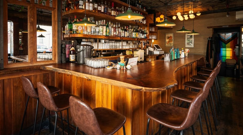Warm, inviting bar with a live-edge wooden counter, brown leather stools, and shelves stocked with bottles.
