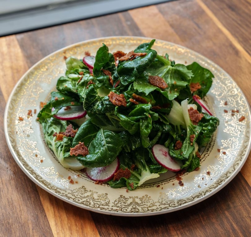 Vibrant green salad with sliced radishes and crispy bacon bits on an ornate plate.