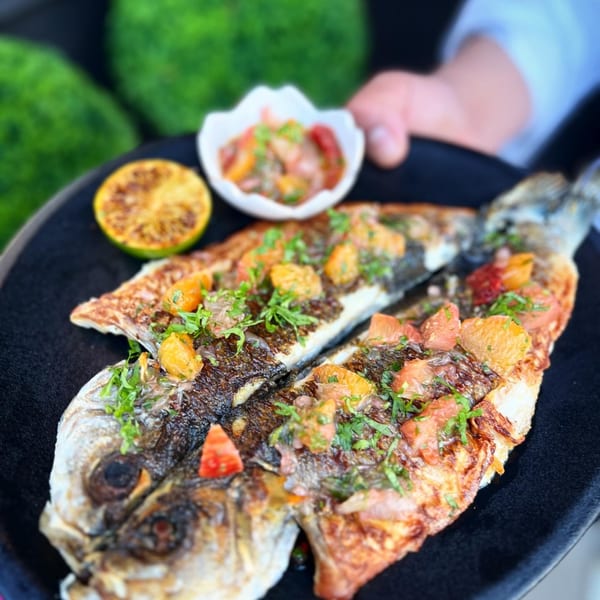 Chef's hand presenting a whole grilled fish topped with citrus, herbs, grilled lime, and salsa.