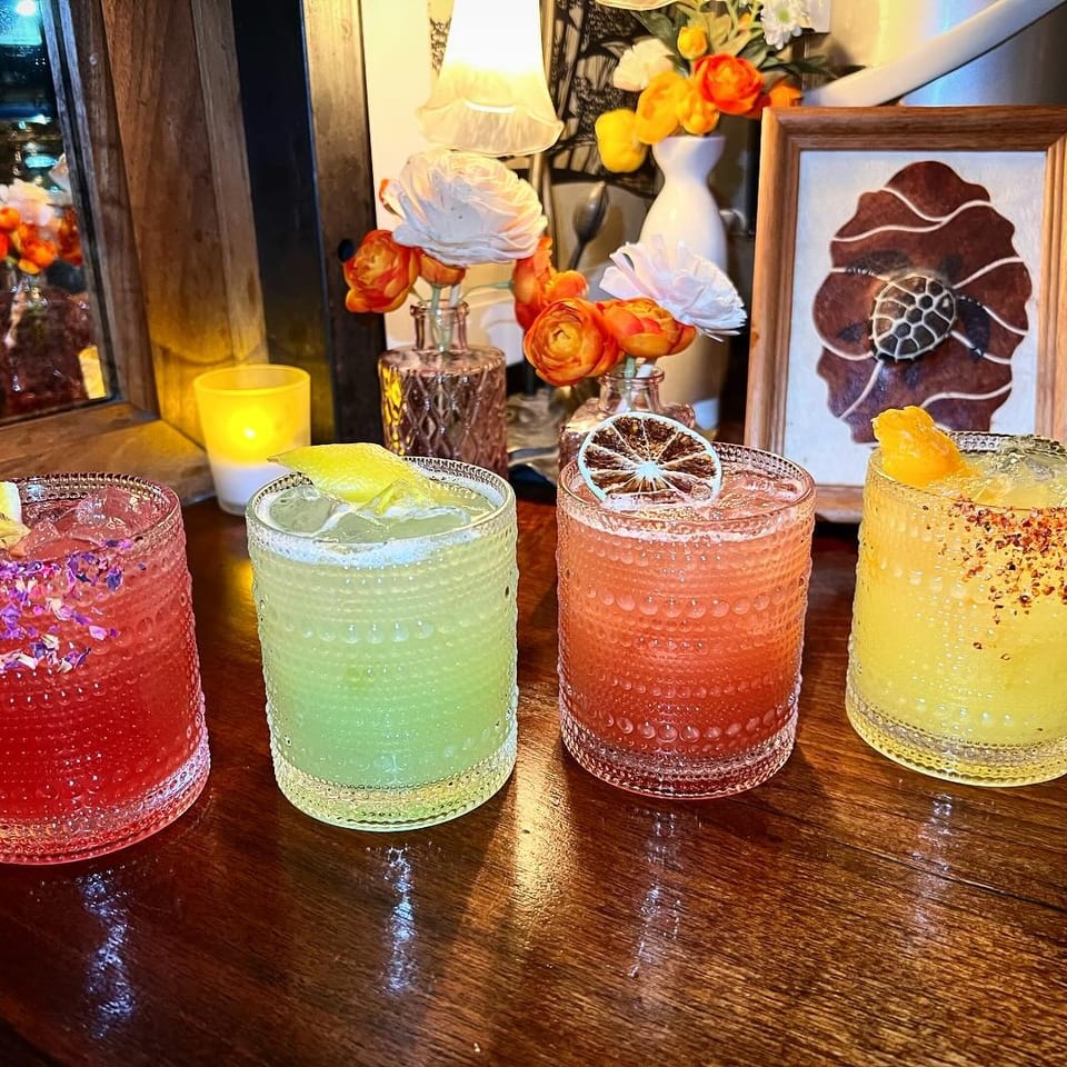 A row of four colorful, garnished cocktails on a dark wooden bar in a dimly lit setting.