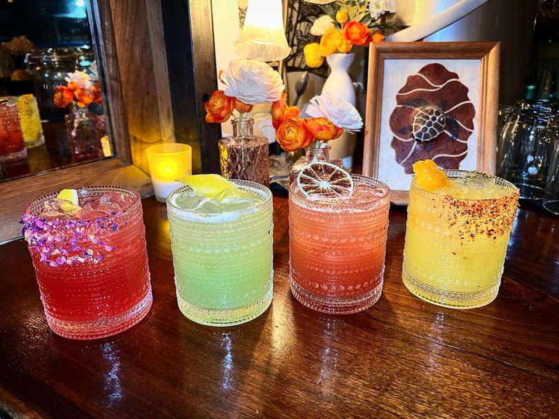 A row of four colorful, garnished cocktails on a dark wooden bar in a dimly lit setting.