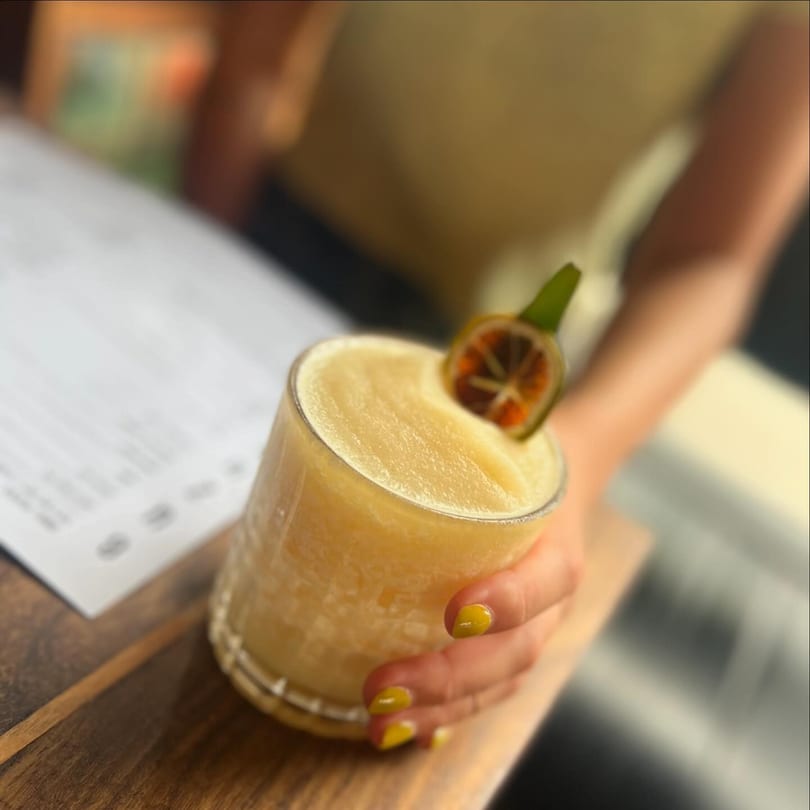 Hand with yellow nails holding a frozen cocktail garnished with a lime on a wooden table.