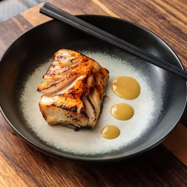 Grilled white fish with a golden-brown seared top, yellow sauce drops, and black chopsticks on a dark plate.
