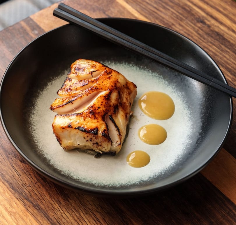 Grilled white fish with a golden-brown seared top, yellow sauce drops, and black chopsticks on a dark plate.