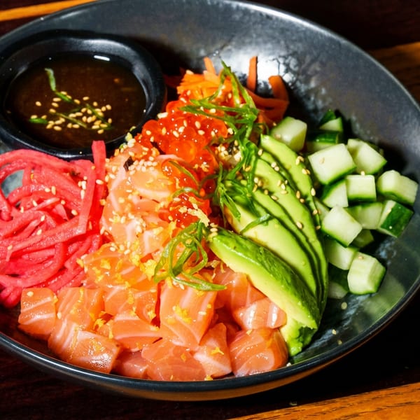 Colorful poke bowl with salmon, avocado, cucumber, red radish, salmon roe, and a side of sauce.