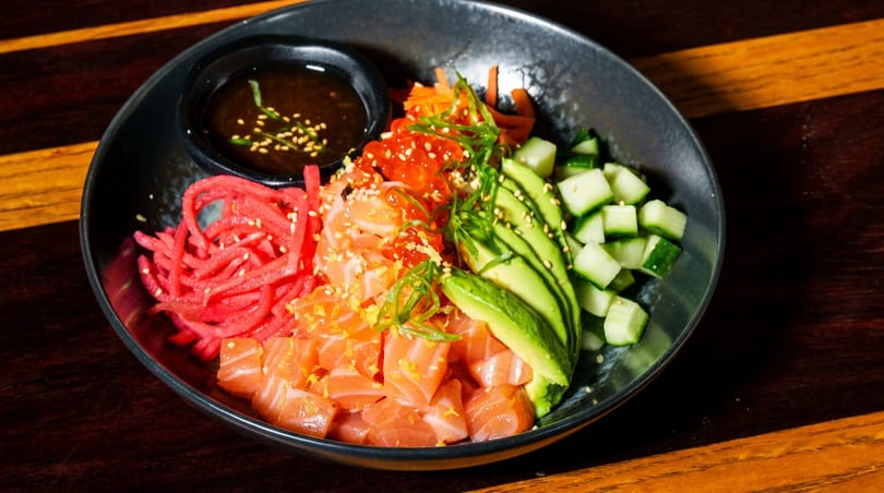 Colorful poke bowl with salmon, avocado, cucumber, red radish, salmon roe, and a side of sauce.